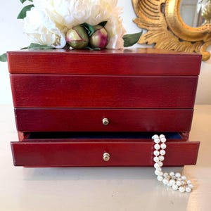 May include: A cherry-red wooden jewelry box with three drawers, one open, revealing a blue interior. A strand of white pearls hangs from the open drawer. Decorative flowers and a gold mirror are in the background.