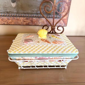 May include: A square ceramic dish with a lid, featuring a yellow and white checkered pattern. The lid is decorated with seashells and a yellow seashell handle. The dish sits in a white wire rack.
