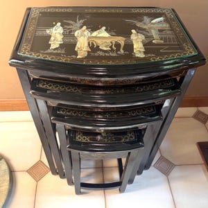 May include: A set of four black nesting tables. The top table features a black surface with a detailed Asian-inspired scene in gold and cream. The tables are stacked, showcasing their graduated sizes and decorative design.