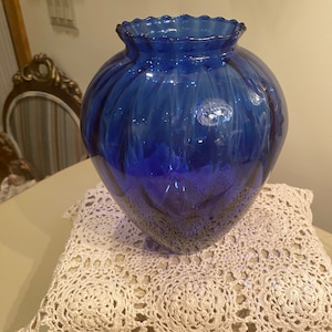 May include: A cobalt blue glass vase with a ruffled top and a ribbed design. The vase is sitting on a white crocheted doily.