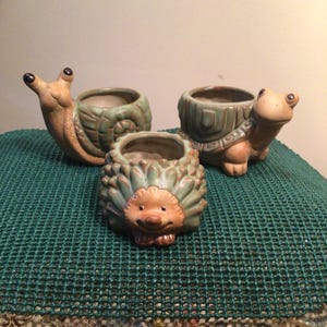 May include: Three ceramic planters shaped like animals: a snail, a hedgehog, and a turtle. The snail is tan and green, the hedgehog is brown and green, and the turtle is tan and green. Each planter has a round opening for planting.