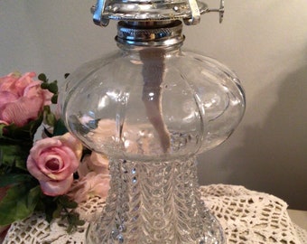 May include: A clear glass oil lamp with a decorative, textured base and a bulbous reservoir. The lamp features a silver-colored metal top with a wick and adjustable levers. The lamp is displayed on a white lace doily, with pink roses in the background.
