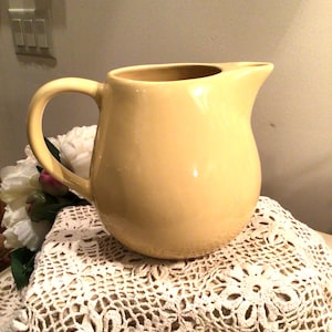 May include: A cream-colored ceramic pitcher with a rounded body, a curved handle, and a spout. The pitcher sits on a white lace doily. The background is a neutral wall with a framed picture and light switches.