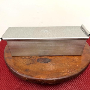 May include: A rectangular, silver-colored loaf pan with a ridged exterior and a rolled handle. The pan is sitting on a round, wooden stool with a dark finish. The background is a plain, off-white wall.