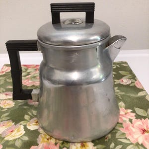 May include: A vintage aluminum coffee percolator with a black handle and lid handle. The coffee pot has a spout and a glass viewing bubble on the lid. The percolator sits on a floral patterned tablecloth with pink and yellow flowers.