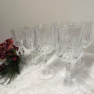 May include: A collection of clear crystal wine glasses with a textured pattern. The glasses have a stem and a round base. The glasses are arranged on a white lace tablecloth. Decorative floral arrangement is visible on the left.