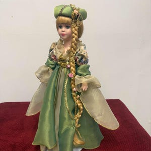 May include: A porcelain doll in a green dress with gold trim and a matching hat. The doll has blonde hair in a long braid adorned with pink roses and gold accents. The doll's sleeves are a sheer, light gold fabric.