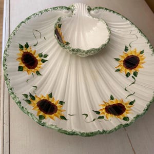 May include: White ceramic serving dish with a sunflower design. The dish has a scalloped edge with a green trim and a smaller, shell-shaped bowl in the center. The sunflowers are yellow and brown with green leaves.