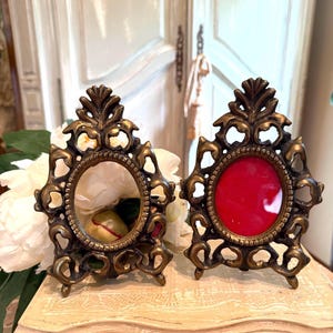 May include: Two ornate, antique-style oval picture frames. The frames are made of bronze-colored metal with intricate scrollwork and a decorative top. One frame has a red background, the other has a white background.