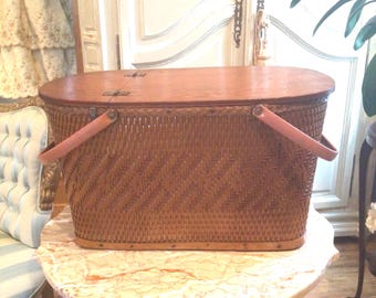 May include: A brown wicker picnic basket with leather handles and a wooden lid. The basket is sitting on a marble table.