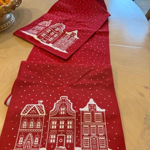 May include: Red Christmas table runner with white dotted pattern and white illustrations of houses. The runner is made of fabric and is folded on a wooden table. The houses have detailed windows and doors.