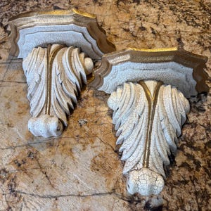 May include: Two ornate, antique-style wall shelves. The shelves are white with gold trim and feature a detailed, leaf-like design. The distressed finish adds to the vintage aesthetic. These decorative shelves are ideal for displaying small items.