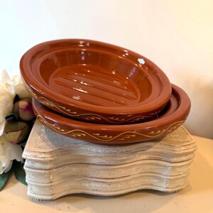 May include: Two terracotta-colored ceramic dishes stacked on a distressed, off-white decorative stand. The top dish has a ribbed interior. The bottom dish has a decorative, cream-colored, wave-like pattern around the rim. The dishes are likely for serving.