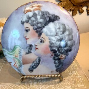 May include: A round, hand-painted porcelain plate featuring two women in profile with elaborate powdered wigs. The plate has a lavender background with gold trim and is displayed on a gold-colored metal stand. The women have rosy cheeks and red lips.