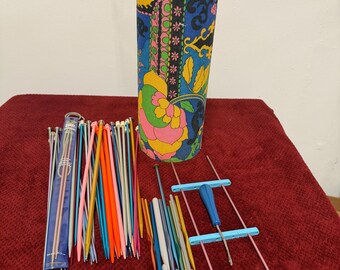 May include: A colorful, cylindrical knitting needle case with a black handle and clasp, filled with various knitting needles and crochet hooks. The case features a vibrant floral pattern in shades of blue, yellow, pink, and green. A variety of knitting tools are arranged on a red surface.