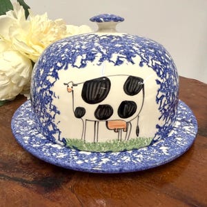 May include: A ceramic butter dish with a blue and white splatter pattern. The lid features a hand-painted cow illustration with black spots and a pink udder. The dish sits on a matching plate, creating a charming farmhouse aesthetic.