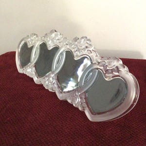 May include: A set of four clear heart-shaped picture frames. The frames are made of a frosted glass material and are decorated with floral accents. The frames are arranged in a row and sit on a burgundy surface.
