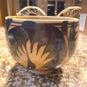 May include: A brown ceramic bowl with a black and brown design of stylized birds. The bowl has a wide rim and a rounded base.
