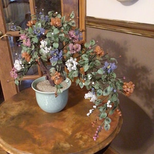 May include: A jade bonsai tree with pink, white, and blue flowers. The tree is potted in a light green ceramic pot and sits on a brown wooden table.
