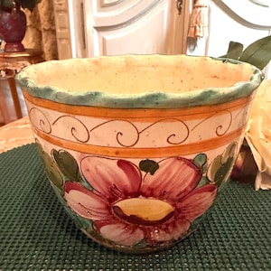 May include: A hand-painted ceramic bowl with a wide, irregular rim. The bowl features a large pink flower design with green leaves and a yellow center. The exterior has a cream-colored base with orange and teal accents.