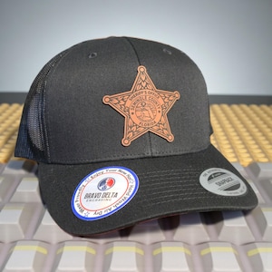 May include: A black trucker hat with a mesh back and a curved brim. The hat features a leather patch with the words "Sheriff's Office Seminole County Florida" and a star design. A Bravo Delta Engraving sticker is on the brim.