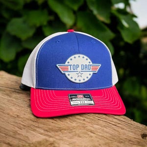May include: A blue, white, and red trucker hat with the words "TOP DAD" in a logo on the front. The hat has a red brim and a white mesh back. The hat is sitting on a wooden surface.