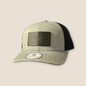 May include: A gray and black trucker hat with a black leather American flag patch on the front. The hat has a black mesh back and a gray brim. The hat has a Brave Delta logo on the side.