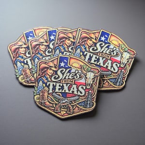 May include: A stack of embroidered patches featuring a woman in a cowboy hat with the text "She's From Texas." The patches have a colorful design with a Texas flag, a longhorn skull, a boot, and a gun.