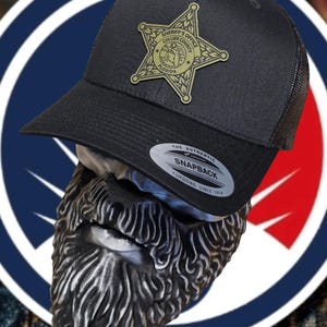 May include: Black mesh trucker hat with a gold star-shaped patch that says "Sheriff's Office Collier County Florida". The hat has a black brim and a black mesh back. The hat has a sticker that says "The Authentic Yupoong Snapback Yupoong Since 1974".