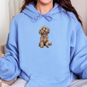 May include: A light blue hooded sweatshirt featuring a graphic of a brown dog centered on the chest. The hoodie has long sleeves, a front kangaroo pocket, and matching blue drawstrings at the neckline. The text CAROLINA BLUE HOODIE is visible.