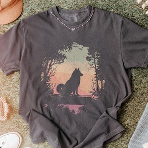 May include: Dark gray t-shirt featuring a wolf silhouette in a forest scene with a sunset backdrop. The shirt has a distressed look and is paired with white sneakers, a pink hat, and a patterned blanket.