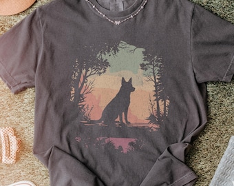 Può includere: T-shirt grigio scuro con la silhouette di un cane in una scena di foresta con uno sfondo di tramonto colorato. La maglietta ha il testo "Pepper T-Shirt". Un cappello rosa, scarpe bianche e una coperta grigia con cuori bianchi sono anche nell'immagine.