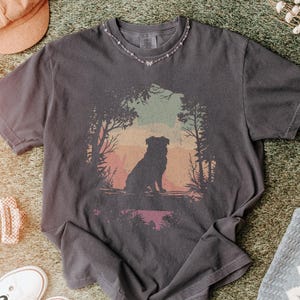May include: Dark gray t-shirt with a graphic of a dog silhouette in a forest scene with a colorful sunset. The shirt has a crew neck and short sleeves. The text "Pepper T-Shirt" is at the bottom left.