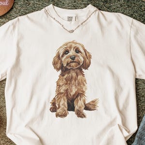 May include: Ivory-colored t-shirt featuring a detailed illustration of a brown dog. The dog is sitting, with its fur rendered in shades of brown. The shirt has a crew neck and short sleeves. The word "ivory" is printed at the bottom of the image.
