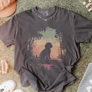 May include: Dark gray t-shirt featuring a silhouette of a dog in a forest scene with colorful sunset hues. The shirt is surrounded by a pink hat, white sneakers, and a light blue blanket with white hearts. The text "PEPPER T-SHIRT" is at the bottom.