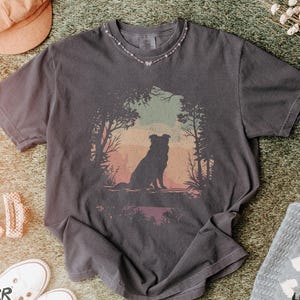 May include: Charcoal grey t-shirt featuring a dog silhouette in a forest setting with a sunset backdrop. The shirt is part of the "Pepper T-Shirt" collection. Accessories include a pink hat, white trainers, and a small necklace.