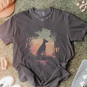 Pu&ograve; includere: T-shirt grigio scuro con una grafica di una silhouette di cane in una scena di foresta con un tramonto colorato. La maglietta &egrave; abbinata a scarpe da ginnastica bianche, un cappello rosa e fermagli per capelli. Il testo "Pepper T-Shirt" &egrave; visibile.