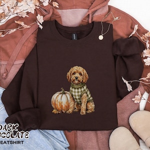 May include: A dark chocolate brown crewneck sweatshirt featuring a graphic illustration of a golden-colored dog wearing a green and white checkered sweater, sitting next to a large, light orange pumpkin. The text DARK CHOCOLATE SWEATSHIRT is visible.