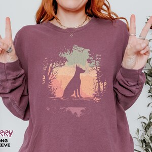 Pu&ograve; includere: Maglietta a maniche lunghe color bacca con la silhouette di un cane in una scena di foresta con sfondo tramonto. La parola "BERRY" &egrave; stampata in basso a sinistra. Il design &egrave; circolare.