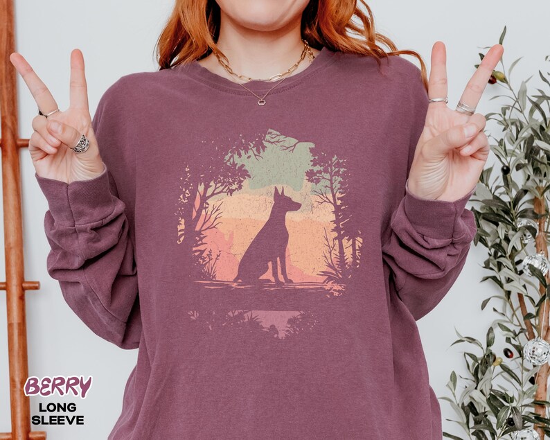 Pu&ograve; includere: Maglietta a maniche lunghe color bacca con la silhouette di un cane in una scena di foresta con sfondo tramonto. La parola "BERRY" &egrave; stampata in basso a sinistra. Il design &egrave; circolare.