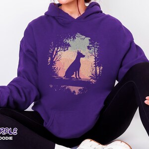 Pu&ograve; includere: Una felpa con cappuccio viola con la silhouette di un cane in una scena di foresta con uno sfondo di tramonto. Il testo "PURPLE HOODIE" &egrave; stampato in basso a sinistra della felpa.