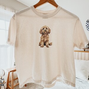 May include: An ivory boxy t-shirt featuring a centered graphic illustration of a brown, fluffy dog sitting down. The shirt has a crew neckline and short sleeves, with the text "IVORY BOXY TEE" printed in the bottom left corner of the image.