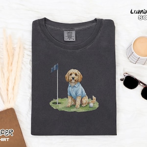May include: Dark gray t-shirt featuring a watercolor illustration of a dog wearing a blue polo shirt, sitting on a golf course with a flag and golf ball. The shirt has the text "Pepper T-Shirt" and the brand name "Luminary Soul".