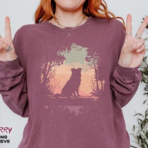 May include: Berry long-sleeve shirt featuring a dog silhouette in a forest scene. The graphic shows a dog sitting before a colourful sunset. The shirt is a deep berry colour.