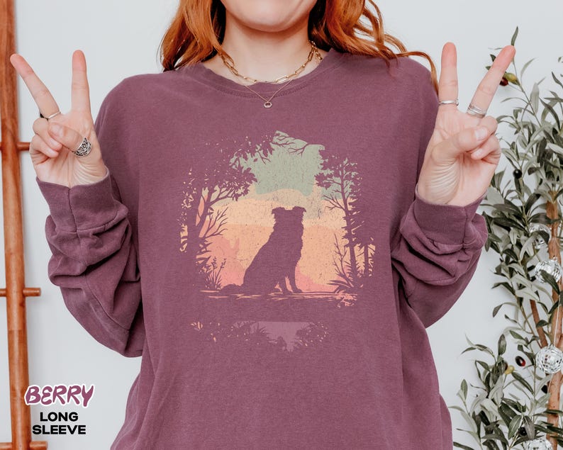 May include: Berry long-sleeve shirt featuring a dog silhouette in a forest scene. The graphic shows a dog sitting before a colourful sunset. The shirt is a deep berry colour.
