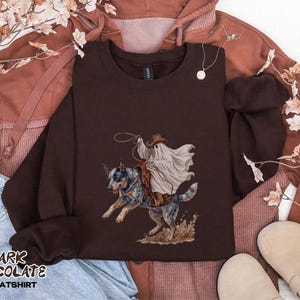 May include: A dark chocolate brown crewneck sweatshirt featuring a graphic print of a ghost wearing a cowboy hat and riding a blue heeler dog. The ghost holds a lasso, and the dog is depicted in motion with dust kicking up at its feet.