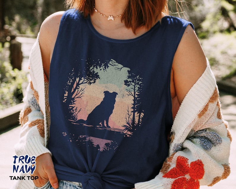 May include: A navy blue vest top featuring a circular graphic of a dog silhouette in a forest scene. The graphic shows a dog sitting, with trees and a colourful sunset background. The text "TRUE NAVY TANK TOP" is printed on the bottom left.