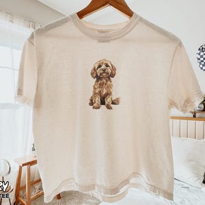 May include: An ivory boxy t-shirt featuring a centered graphic illustration of a brown, fluffy dog sitting down. The shirt has a crew neckline and short sleeves, with the text "IVORY BOXY TEE" printed in the bottom left corner of the image.