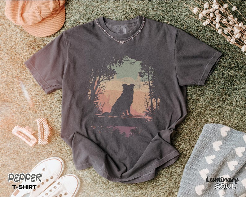 May include: Charcoal grey t-shirt featuring a dog silhouette in a forest setting with a sunset backdrop. The shirt is part of the "Pepper T-Shirt" collection. Accessories include a pink hat, white trainers, and a small necklace.