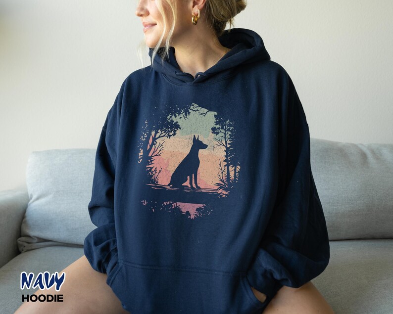 Pu&ograve; includere: Felpa con cappuccio blu navy con la silhouette di un cane in una scena di foresta. Il design presenta una sfumatura di colore rosa, arancione e verde. Le parole "NAVY" e "HOODIE" sono stampate in basso a sinistra dell'immagine.